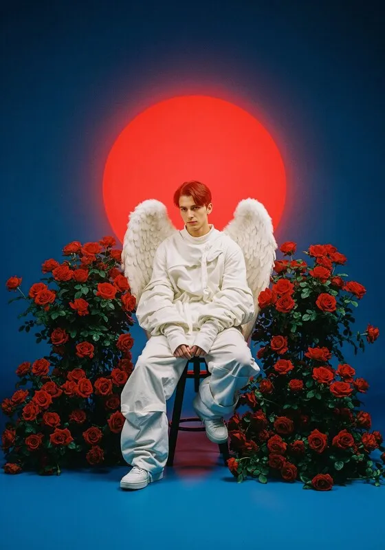 A photograph of a young man with bright red hair, wearing a white oversized jumpsuit and white angel wings. He is sitting on a chair between two large bushes of vibrant red roses. In the background, there is a bold, red sun-like sphere against a deep blue sky. The overall mood is stylized and a bit melancholic.