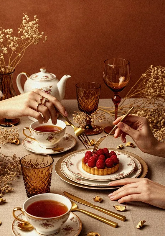A photograph of a high-end tea time setting with two people's hands visible. One hand is holding a fork to eat a raspberry tart on a decorative plate, while the other is holding a spoon in a teacup. The table is set with delicate teacups, a teapot, elegant glassware, and golden cutlery. The background is a solid brown color with dried flowers, creating a warm and sophisticated mood.